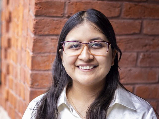 Headshot of a student with long dark hair and glasses against a red brick background