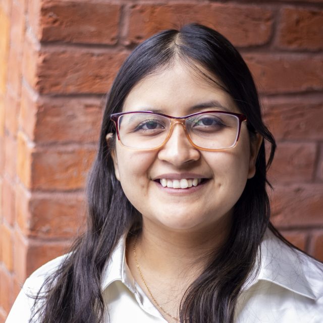 Headshot of a student with long dark hair and glasses against a red brick background